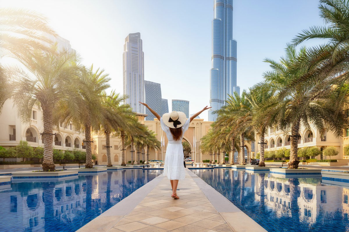 Moving from Melbourne to Dubai Made Easy Woman in a white dress stands by a pool, with palm trees and the Dubai skyline visible in the background; Moving from Melbourne to Dubai Made Easy