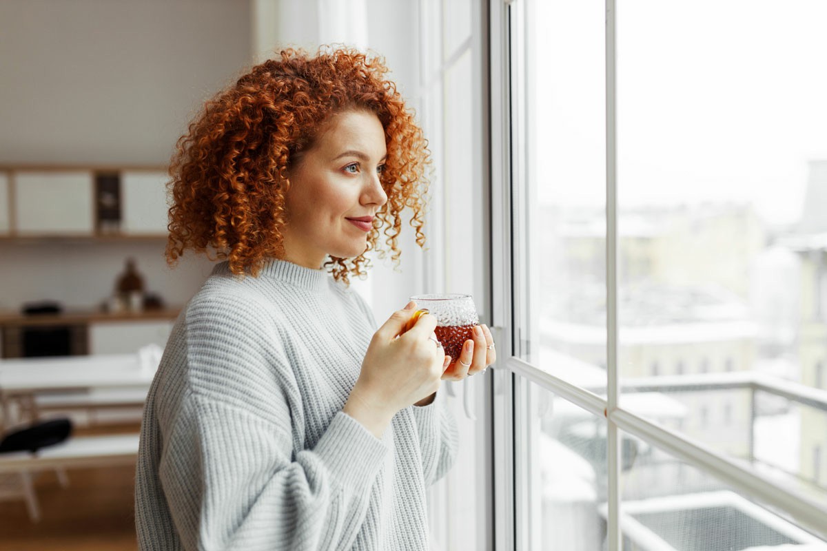 A woman with red curly hair is holding a steaming cup of tea, smiling gently as she enjoys her drink. Moving Balancing Your New Perspective with Your Old Life 