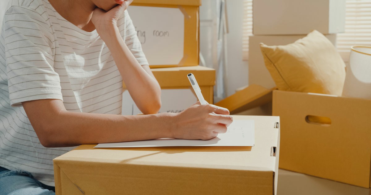 How To Create A Moving Schedule To Simplify Your Move A woman focused at a table, jotting down ideas with a pen and paper to simplify her moving process. How To Create A Moving Schedule To Simplify Your Move
